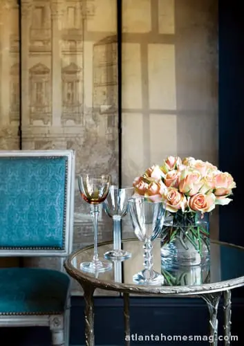 A teal upholstered chair sits next to a small mirrored table holding three wine glasses and a vase of peach roses. Behind the table is a screen with a sketch of buildings.