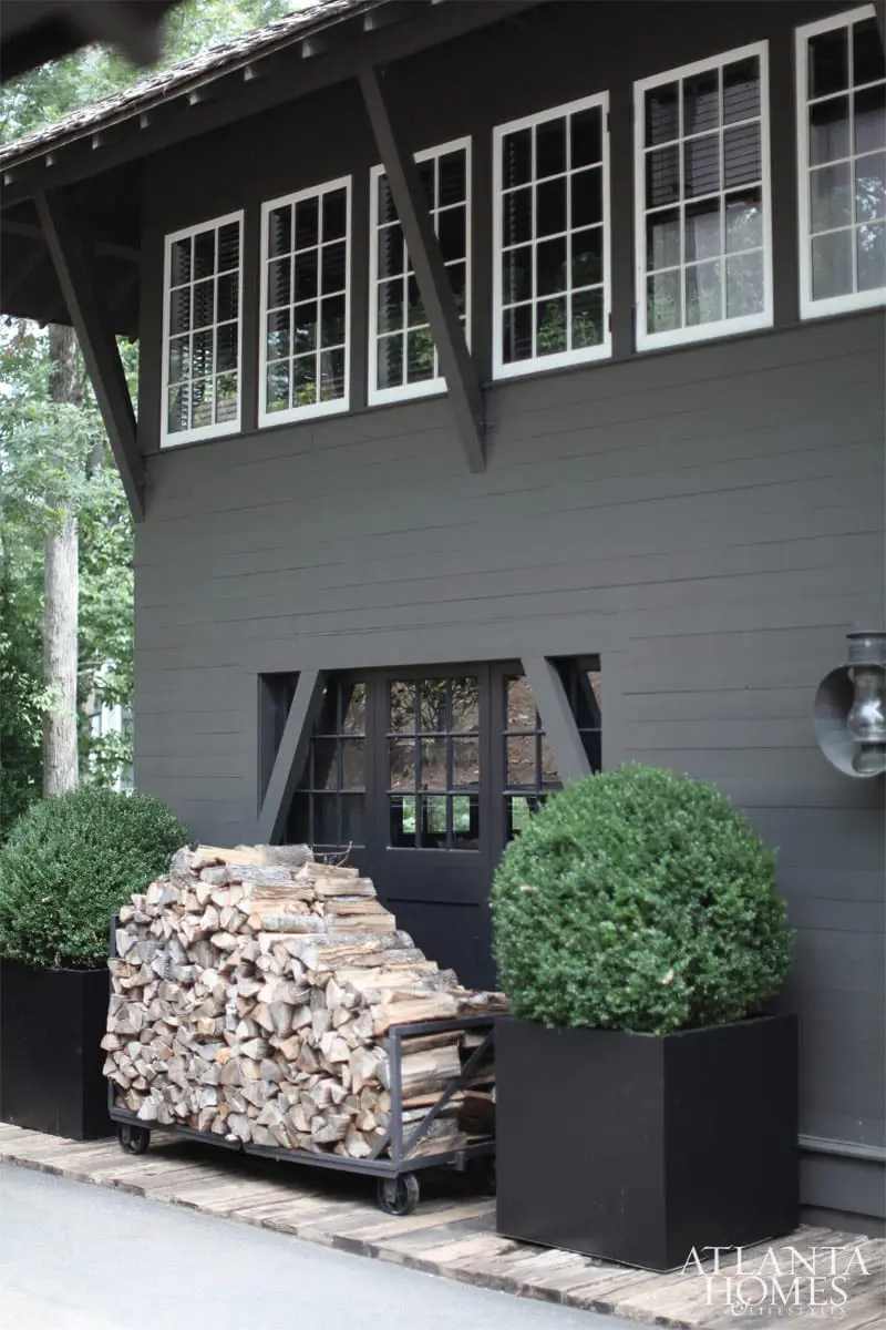 Dark gray wood-sided building with white-paned windows, a dark door with a geometric design, a cart piled with firewood, and two black planters with boxwood shrubs.