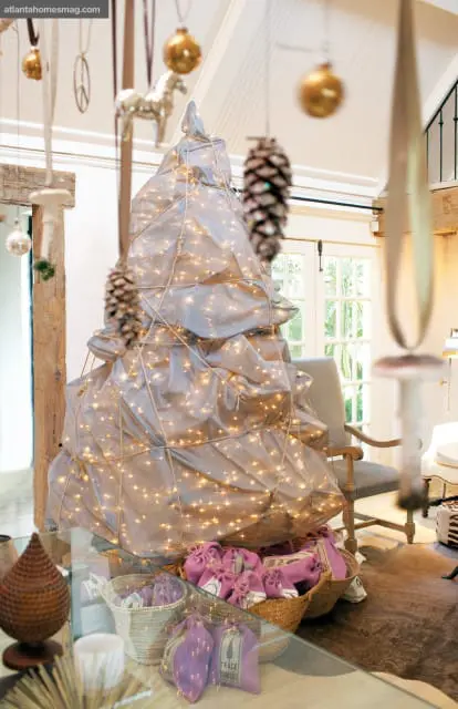 A unique Christmas tree draped in silver fabric and illuminated by fairy lights, adorned with pinecones and ornaments. Baskets of purple gift bags sit at its base.
