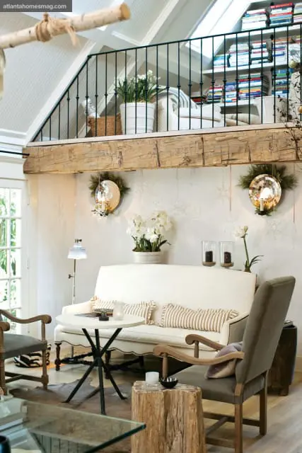 Living room with a white sofa, striped pillows, and a round white table. A rustic wooden beam spans the ceiling above, with a loft area featuring a railing and bookshelves.