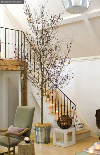 Interior view of a home with a bare tree decorated with gold, pink, and green ornaments in a galvanized bucket. A staircase with black railing and lit candles ascends to the right.