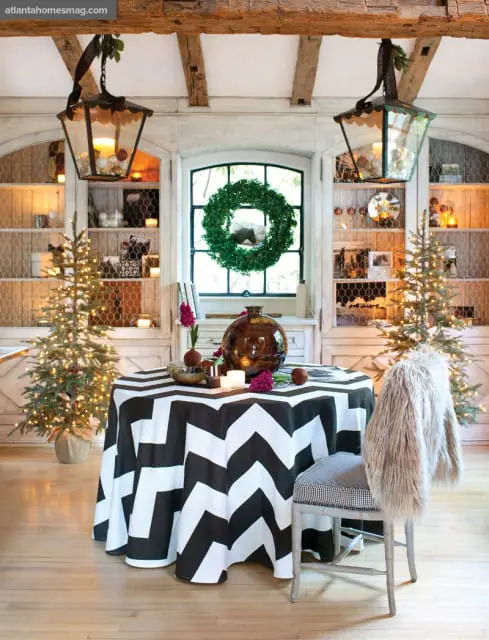 Dining room with a round table covered in a black and white chevron tablecloth, two decorated Christmas trees, and a chair draped with a faux fur throw.