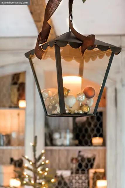 Close-up of a dark metal lantern filled with a large white candle and small gold and silver Christmas ornaments, hanging by a brown ribbon.