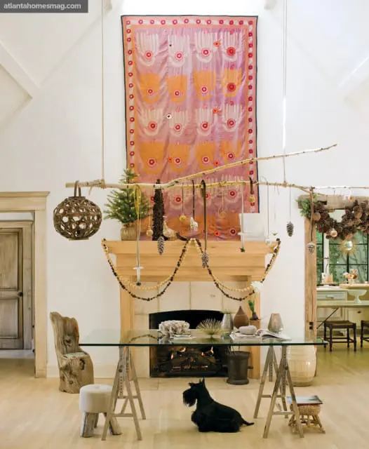 Living room with a stone fireplace, a glass desk, and a Scottish Terrier. A large tapestry hangs on the wall above the mantel, decorated with a small Christmas tree and pinecone garlands.