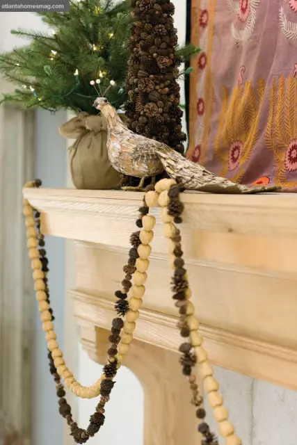 Wooden mantelpiece decorated with a festive garland of nuts and pinecones, a decorative bird, a small Christmas tree with lights, and a patterned textile.