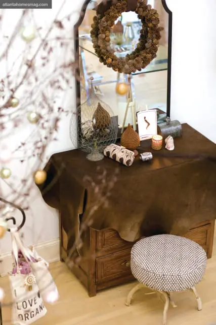 Wooden dresser with a brown leather draped over it, topped with decorative items including a pinecone wreath, a wire birdcage, and a small candle. A patterned stool sits to the right.