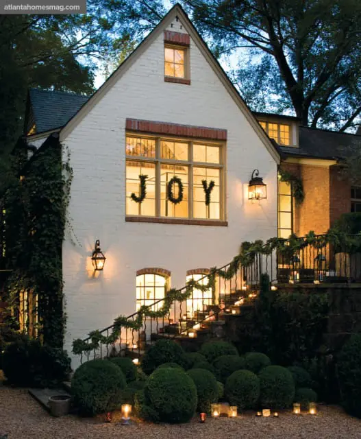 White brick house with a peaked roof, decorated for the holidays with the word "JOY" in the window and garlands and candles lining the exterior staircase.