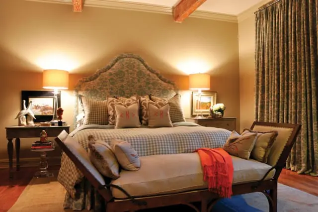 Bedroom with a large upholstered headboard in a floral pattern, gingham bedding, and decorative pillows. A bench with an orange throw sits at the foot of the bed.