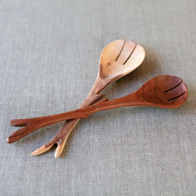 Two wooden salad servers, one lighter wood and one darker wood, with carved tines and a textured linen background.
