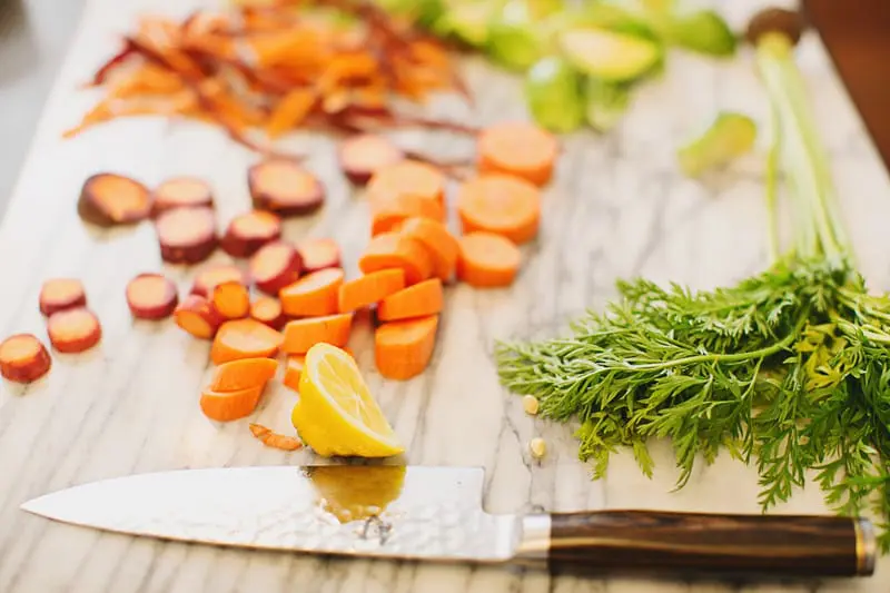 Chopped carrots and purple carrots on a marble cutting board next to a knife, a lemon wedge, and fresh carrot greens.