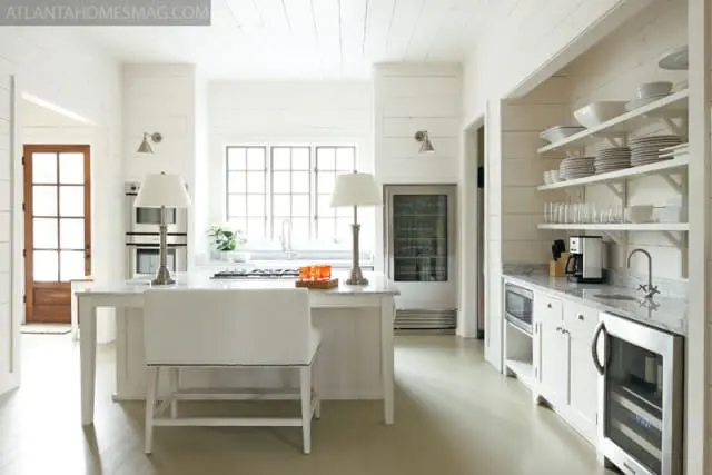 White shiplap kitchen with marble countertops, a central island with a white upholstered stool, stainless steel appliances including a wine fridge and coffee maker, and open shelving displaying dishes.