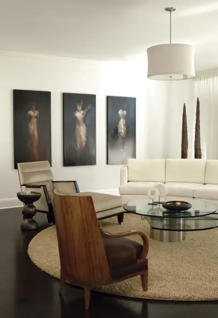 Living room with three abstract paintings of figures on the wall, a cream sofa, a glass coffee table, and two armchairs on a round cream rug.