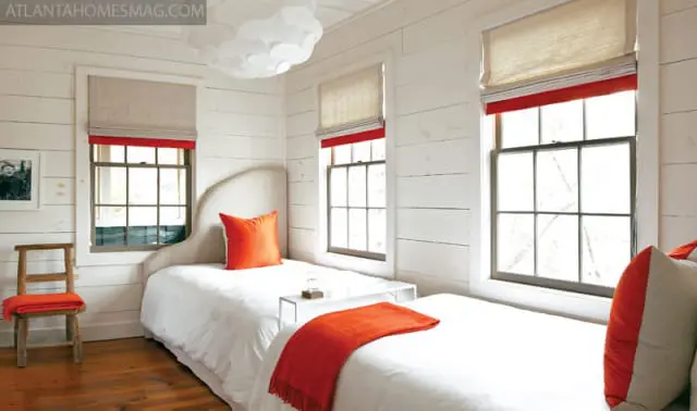Bedroom with white shiplap walls, two twin beds with white linens and orange accents, and windows with Roman shades.
