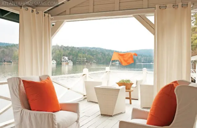 Covered porch with white slipcovered chairs and orange pillows overlooking a lake with houses and trees on the far shore.