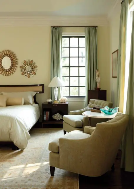 Bedroom with pale yellow walls, a queen bed with white patterned bedding, two sunburst mirrors, a dark wood nightstand with a glass lamp, and two upholstered chairs next to a round marble-top table.