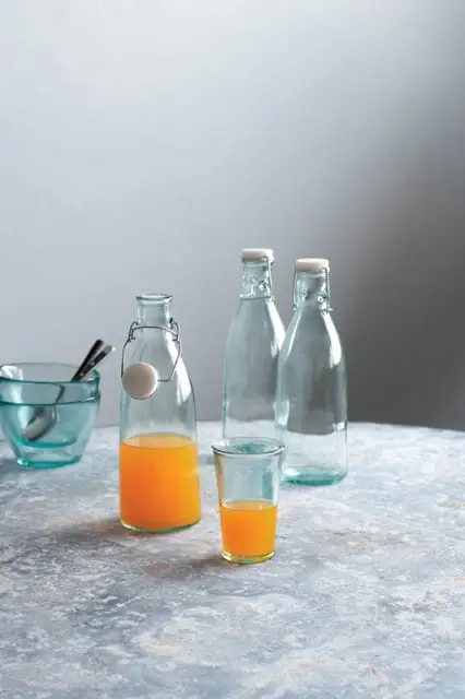 Glass bottle with orange juice and a glass of orange juice, next to two empty glass bottles and a blue glass bowl with spoons on a textured surface.