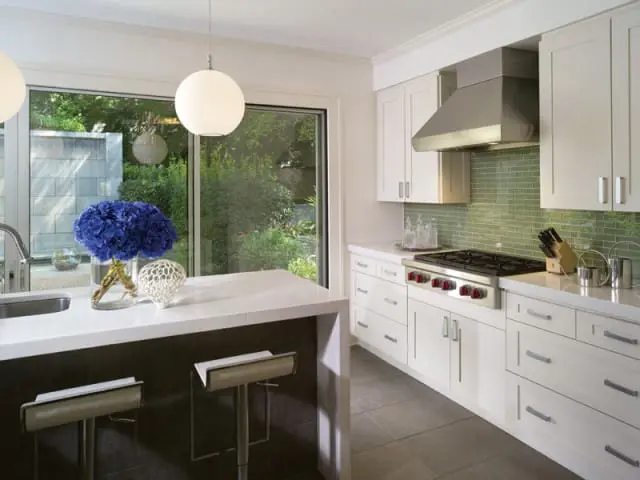 White kitchen with white cabinetry, stainless steel range hood, green subway tile backsplash, and a white quartz island with two bar stools.