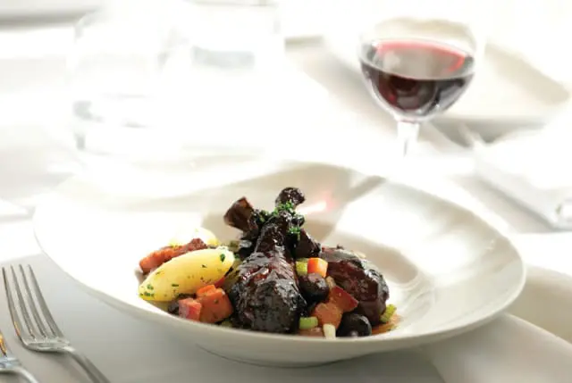Braised duck legs with potatoes, carrots, celery, and bacon in a white bowl, with a glass of red wine and a water glass in the background.