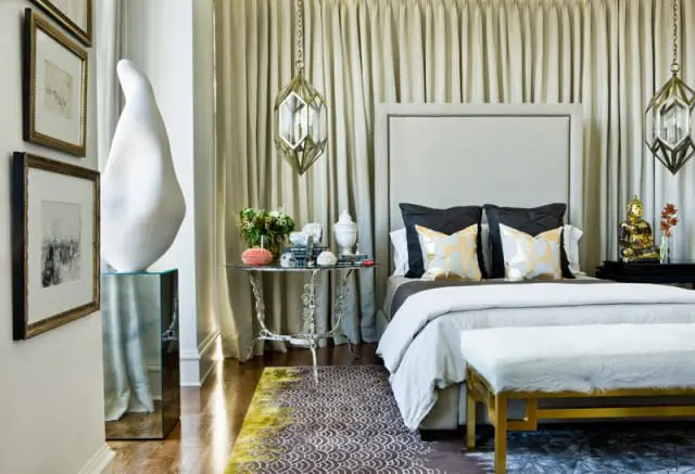 Bedroom with upholstered headboard, dark gray and gold patterned pillows, white duvet, and a white fur bench with gold legs. A gold Buddha statue sits on a black nightstand.