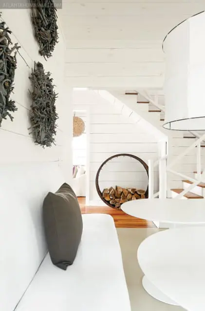 White living room with white sofa, dark gray pillow, white tables, and a circular wood rack filled with logs next to a staircase.