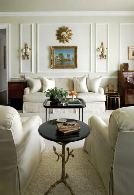 Living room with a cream slipcovered sofa, two cream armchairs, and a black coffee table with books and a plant. Walls are pale green with decorative molding and gold sconces.