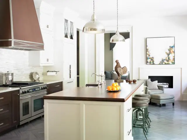 White kitchen island with wood countertop, dark wood lower cabinets, stainless steel range with copper hood, and a fireplace with armchair in the background.