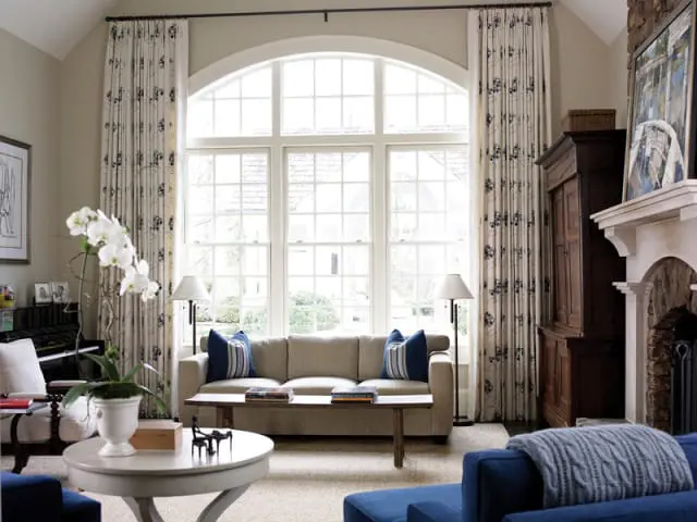 Living room with a large arched window, beige sofa with blue pillows, floral curtains, a dark wood cabinet, and a stone fireplace.