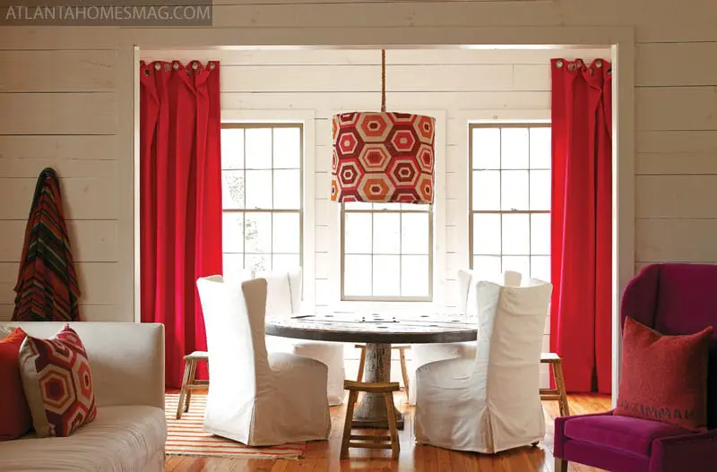 Dining area with white slipcovered chairs around a round table, red curtains, and a hexagonal patterned lampshade.