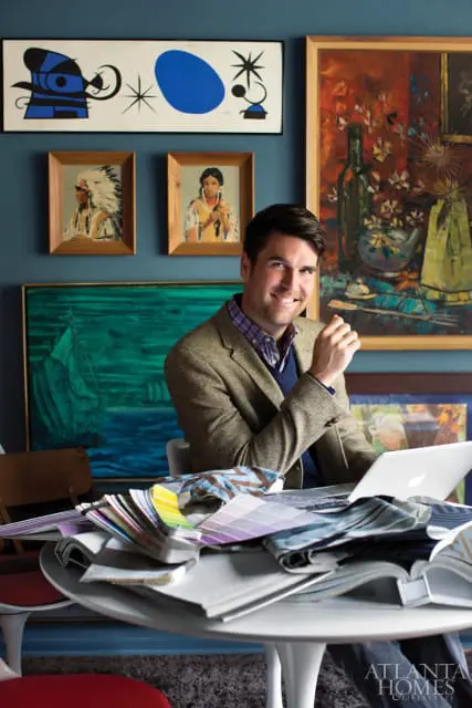 Man in tweed jacket sitting at a table with fabric swatches and a laptop, surrounded by colorful artwork on a blue wall.