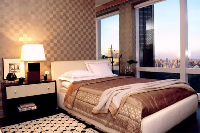 Bedroom with a beige upholstered bed, patterned brown bedding, and a large window overlooking a city skyline at dusk. A nightstand with a lamp and artwork sits beside the bed.