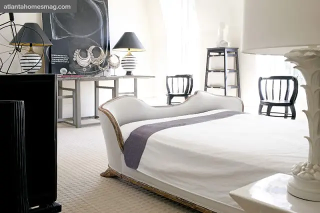 Bedroom with a white chaise lounge, black and white striped lamps, a metal console table with artwork and decorative necklaces, and two black chairs.