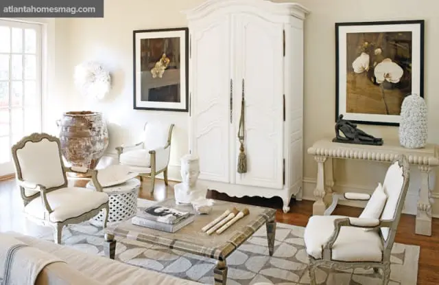 Living room with a large white armoire, upholstered chairs, a patterned rug, and decorative art pieces including a large urn and framed floral prints.