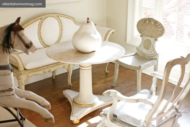 Ornate antique white settee and chairs surround a marble-top pedestal table with a pear sculpture in a sunlit room.