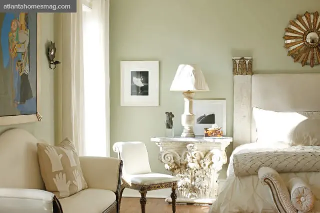 Bedroom with pale green walls, featuring a cream upholstered headboard, a white duvet, and a textured throw blanket. A carved pedestal table holds a lamp and framed art.