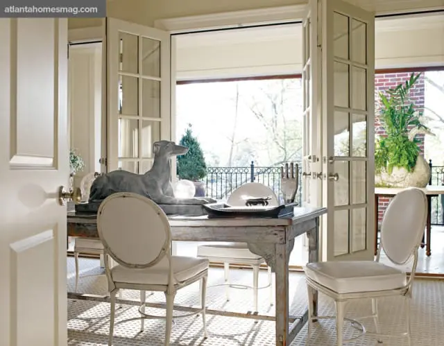 Dining room with a distressed wood table featuring a grey dog sculpture and white upholstered chairs, opening to a sunlit balcony with greenery.