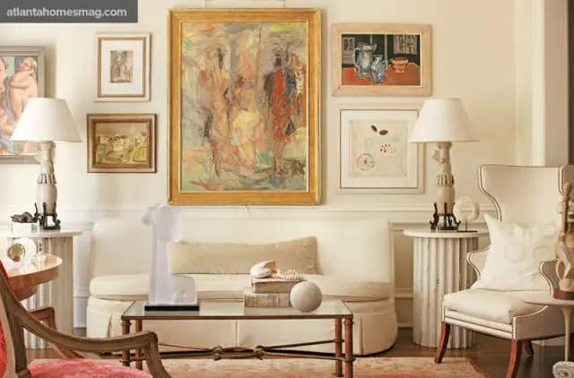 Living room with a white sofa, glass coffee table, and multiple framed artworks on the wall. A wingback chair sits to the right.