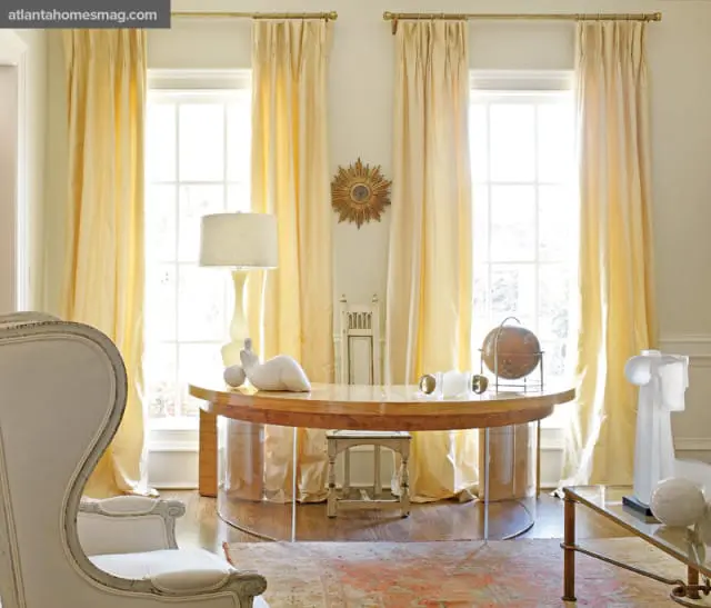Elegant home office with a curved wooden desk, a white wingback chair, and tall yellow drapes framing large windows. A globe and sculptural art pieces adorn the space.