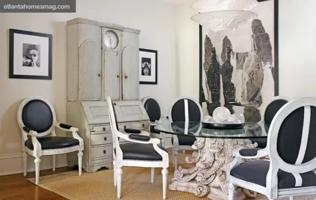 Dining room with a distressed antique secretary desk, black and white upholstered chairs, and a glass-top table with a carved pedestal base.