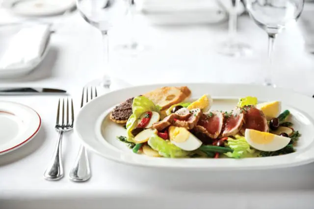 Seared tuna salad with hard-boiled eggs, olives, green beans, and toast points on a white plate, set on a white tablecloth with silverware and wine glasses.