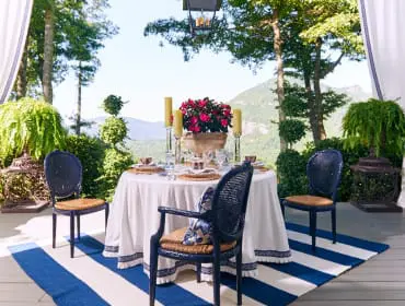 Outdoor dining area with a round table set for four, navy cane-back chairs, and a blue and white striped rug, overlooking a mountain landscape.