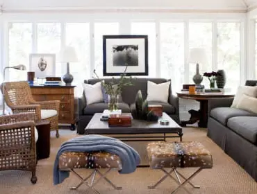 Living room with two dark gray sofas, wicker armchairs, a dark wood coffee table, and two animal print ottomans. Large windows provide natural light.
