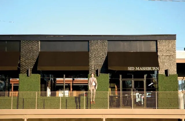 Man in a suit standing on a balcony in front of a building with black awnings that read "SID MASHBURN"