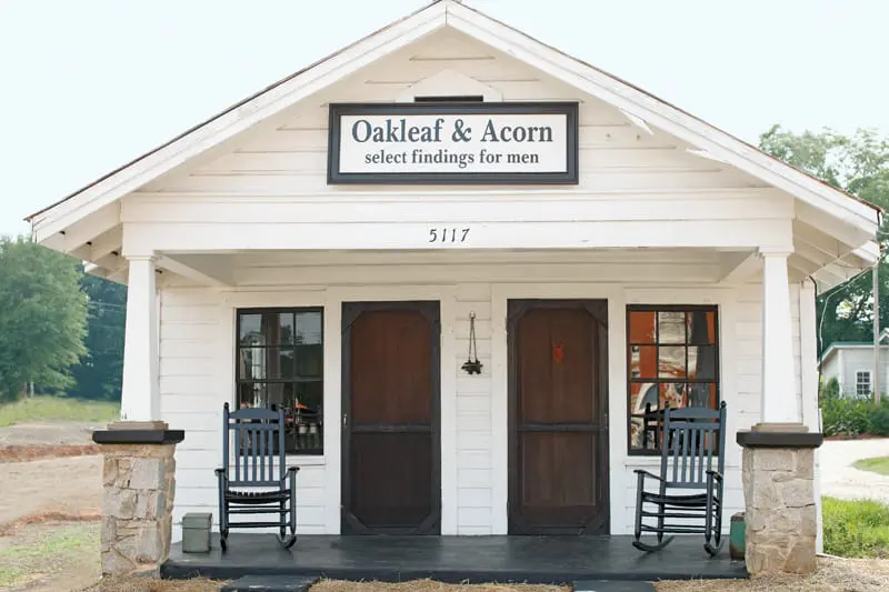 White clapboard building with a gable roof, two dark wooden screen doors, and two rocking chairs on the porch. A sign reads "Oakleaf & Acorn select findings for men".