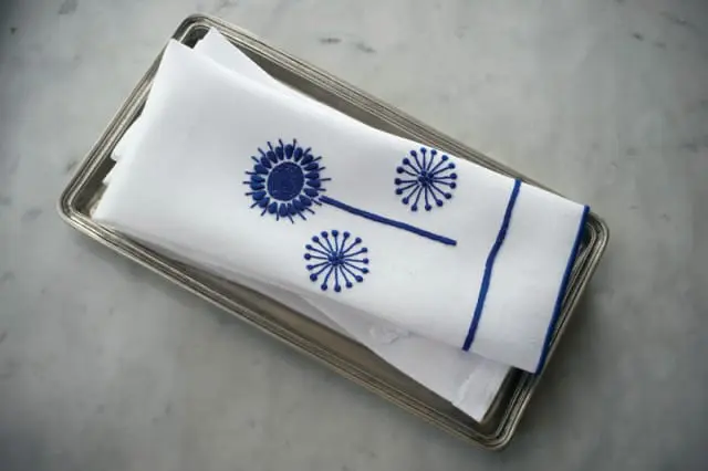 White linen napkins with blue embroidered dandelion motifs and blue border trim, resting on a silver tray.