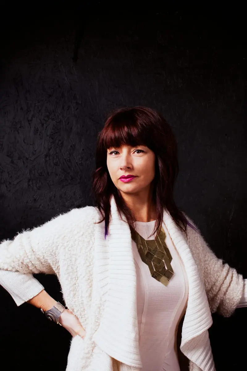 Woman with dark red hair and bangs, wearing a cream-colored textured cardigan over a cream dress and a geometric gold necklace. She has bright pink lipstick and a large silver bracelet on her left wrist.