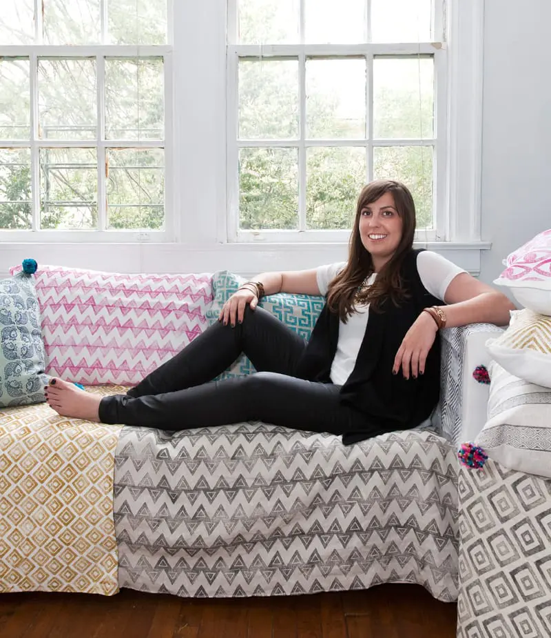 Woman sitting on a sofa upholstered in patterned fabrics with geometric designs in pink, teal, gold, and gray.