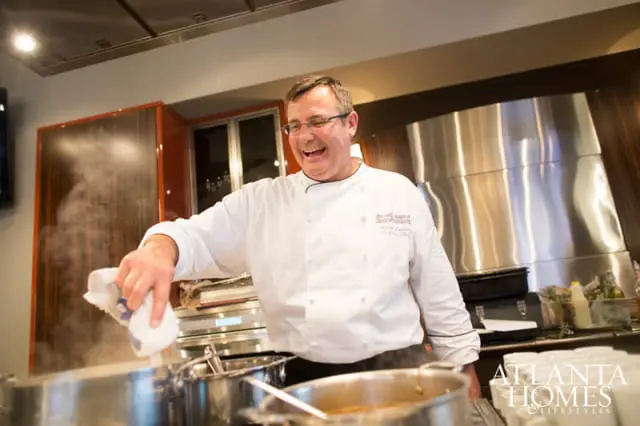 Chef in a white uniform smiles while cooking in a modern kitchen with stainless steel appliances and wood cabinetry.