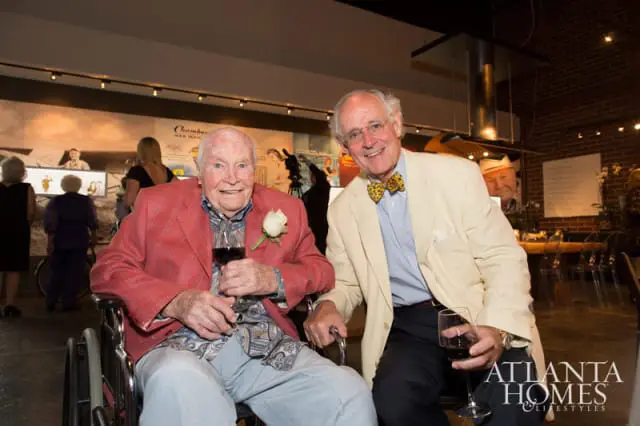 Two men smiling, one in a wheelchair wearing a red jacket and a white rose, the other in a cream jacket and a leopard-print bow tie.