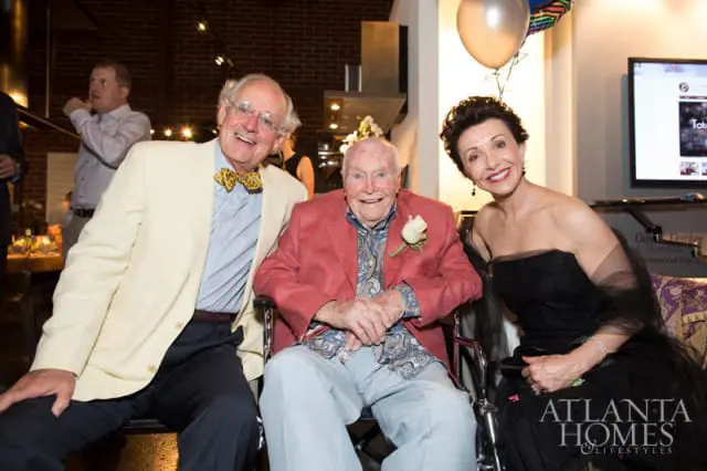 Three smiling people at an event: a man in a cream jacket and yellow bow tie, a man in a wheelchair wearing a red jacket and patterned shirt, and a woman in a black strapless gown.