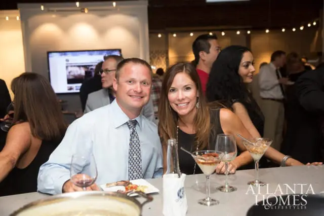 A man in a light blue dress shirt and patterned tie smiles next to a woman in a black dress at a bar. Behind them, a large screen displays content, and other guests mingle.